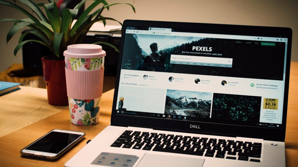 A website homepage is displayed on a laptop, which sits on a wooden desk next to a green plant and a pink and floral reusable coffee cup.